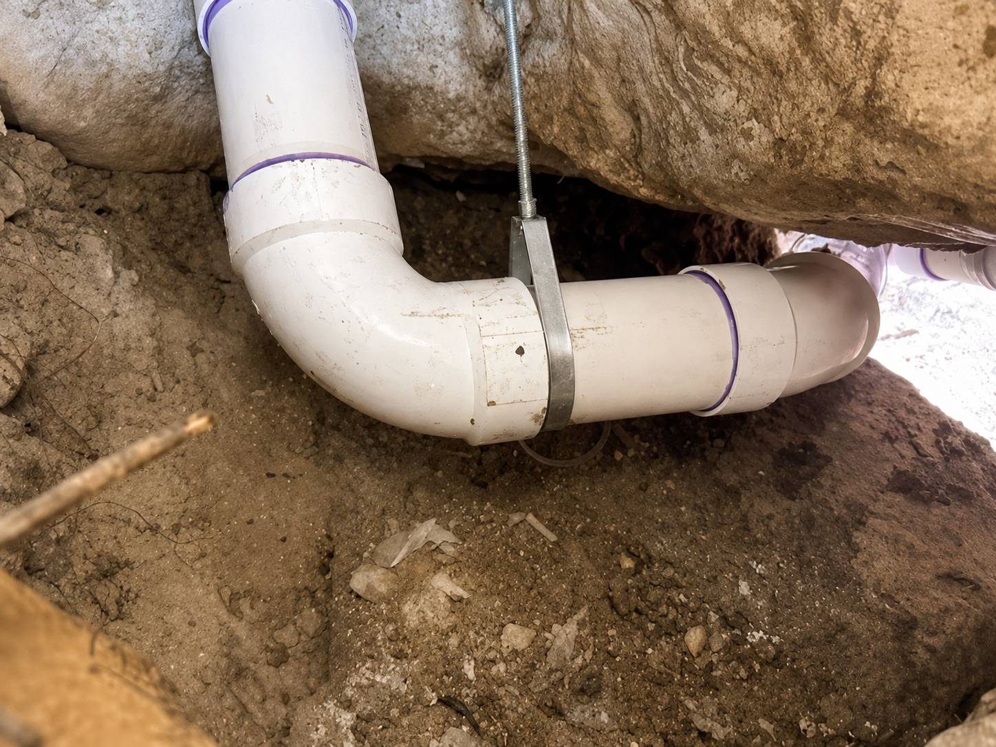 PVC sewer pipe repair beneath a residential foundation, supported by a pipe hanger in compacted soil